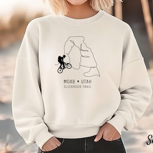 May include: Sand-colored sweatshirt featuring a black line drawing of a cyclist on a mountain bike near a rock formation. The text "MOAB - UTAH SLICKROCK TRAIL" is printed below the graphic. The sweatshirt is a neutral color.