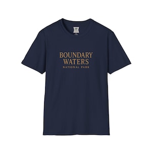 Puede incluir: Camiseta azul marino con el texto "BOUNDARY WATERS NATIONAL PARK" en color dorado. La camiseta tiene cuello redondo y mangas cortas. La tela parece suave.
