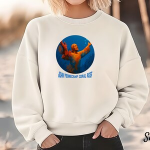 Op de afbeelding: Crèmekleurige sweatshirt met een cirkelvormige afbeelding van een persoon met uitgestrekte armen in een onderwaterlandschap. De afbeelding is omlijst door een blauwe cirkel en bevat de tekst "JOHN PENNECAMP CORAL REEF".