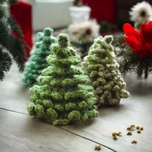 May include: Close-up of several small, handmade crochet Christmas trees in varying shades of green. The trees are arranged on a light-colored wooden surface, with a few small gold-colored star-shaped decorations scattered around them. The trees have a textured, fuzzy appearance.