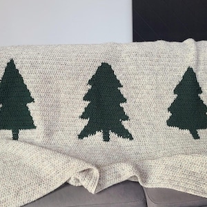 May include: A cream-colored crocheted blanket featuring three dark green Christmas tree designs. The blanket is draped over a gray sofa, showcasing the textured stitch pattern and the contrasting colors of the trees.