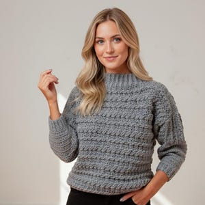 May include: A grey, textured crochet jumper with a high neckline and long sleeves. The jumper has a ribbed collar and cuffs. The model is wearing black trousers. The jumper is a medium weight and appears to be handmade.