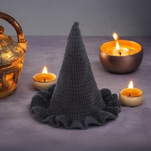 May include: A dark gray crocheted witch hat with a ruffled brim. The hat is cone-shaped and sits on a purple surface, surrounded by lit candles in gold and cream-colored holders. A decorative gold lantern is in the background.