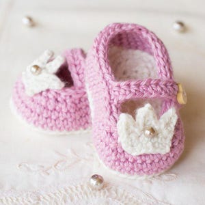 May include: A pair of pink crocheted baby shoes with white crocheted crown embellishments and pearl accents.