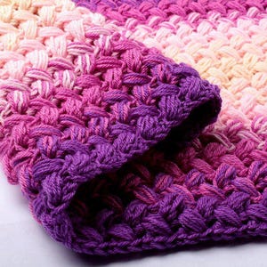 May include: A crocheted blanket with a gradient of pink, purple, and white yarn. The blanket is made with a simple stitch pattern and has a soft, textured feel.