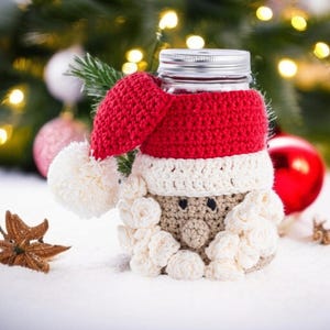 May include: A glass jar with a silver lid, decorated with a crocheted Santa Claus design. The Santa hat is red with a white pom-pom, and the face is beige with white beard and mustache details. Christmas-themed.