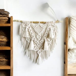 May include: A white macrame wall hanging with a geometric pattern and tassels. The hanging is attached to a wooden dowel rod.