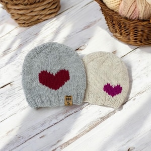 以下が含まれることがあります： ハートのデザインが施されたニット帽が2つ。グレーの帽子には赤いハート、ベージュの帽子には紫のハートがデザインされています。