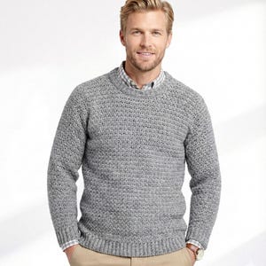 Può includere: Un maglione grigio a maglia testurizzata con girocollo e maniche lunghe. Il maglione è indossato sopra una camicia con colletto. La persona indossa un orologio e pantaloni color cachi. Il maglione è un capo classico e versatile.