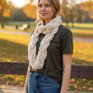 Puede incluir: Una bufanda infinita de punto grueso de color crema. La bufanda está hecha de hilo grueso y tiene un diseño trenzado. La persona de la imagen lleva una camiseta verde oscuro y vaqueros azules. El fondo es un parque con follaje otoñal.