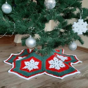 以下が含まれることがあります： 手作りのクリスマスツリースカート。星型のデザインで、赤、緑、白の糸でかぎ針編みされ、雪の結晶模様が特徴です。銀色のオーナメントで飾られたクリスマスツリーの下に置かれています。