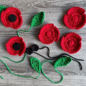 May include: A set of red and green crocheted poppy flowers and leaves. The flowers have black centres and are arranged on a wooden surface.