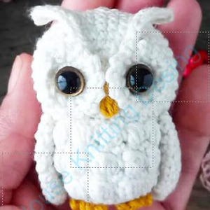 Könnte beinhalten: Eine kleine, weiß gehäkelte Eulenfigur mit großen schwarzen Augen und einem gelben Schnabel und Füßen. Die Eule wird in einer Hand gehalten, wodurch die handgefertigten Details und die Textur des Garns hervorgehoben werden.