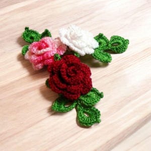 Puede incluir: Tres rosas de ganchillo en rosa, blanco y rojo con hojas verdes. Las rosas están dispuestas en un racimo y están hechas con un punto de ganchillo delicado.