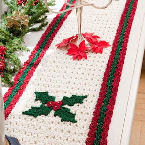 May include: A cream-coloured, crocheted Christmas table runner with a holly leaf design. The runner has a red and green border and is decorated with red poinsettias. A silver candelabra with white candles is on the table.
