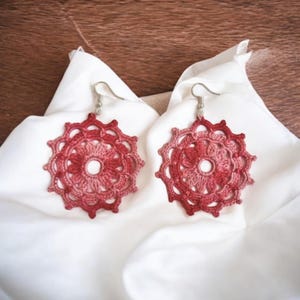 May include: A pair of crocheted earrings with a pink and red ombre design. The earrings are round and have a delicate floral pattern.