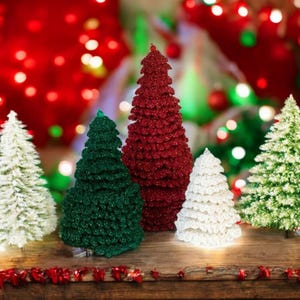May include: A collection of decorative Christmas trees in various sizes and colors. The trees are covered in glitter and range in color from white and green to red. They are arranged on a wooden surface with a blurred background of Christmas lights.
