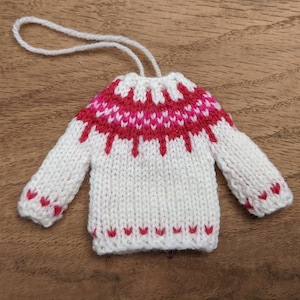May include: White knitted sweater ornament with a red and pink patterned yoke. The ornament has a white string loop for hanging.