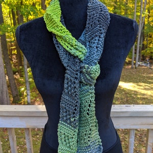 May include: A green and gray knitted scarf with a lacy pattern. The scarf is tied in a knot and draped over a black mannequin.