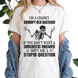 May include: A white short-sleeved t-shirt featuring a black graphic of a skeleton with arms raised. The text reads: I'M A CRANKY GRUMPY OLD BASTARD IF YOU DON'T WANT A SARCASTIC ANSWER DON'T ASK A STUPID QUESTION. The shirt is worn under a blue denim jacket.