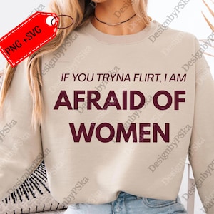 May include: A beige sweatshirt with the text "IF YOU TRYNA FLIRT, I AM AFRAID OF WOMEN" in dark red. The image also includes a red tag with "PNG + SVG" on it. The sweatshirt is a casual style.
