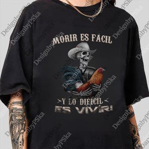 May include: Black t-shirt featuring a skeleton wearing a cowboy hat and holding a rooster. The text "MORIR ES FACIL" and "Y LO DIFICIL ES VIVIR!" are printed above and below the illustration. The design is detailed and colorful.