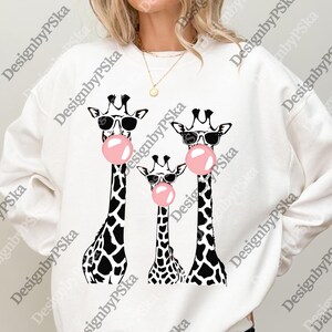 May include: White sweatshirt with a graphic of three giraffes wearing sunglasses and blowing pink bubblegum bubbles. The giraffes have black and white spotted patterns. The text "DesignbyPSka" is printed on the sweatshirt.