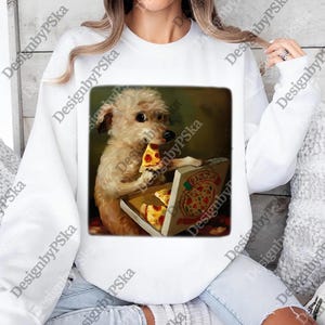 Puede incluir: Sudadera blanca con un dise&ntilde;o cuadrado de un perro peque&ntilde;o comiendo pizza de una caja. La caja de pizza est&aacute; abierta, mostrando porciones de pizza. El perro es de color marr&oacute;n claro y tiene pelaje esponjoso. La sudadera tiene mangas largas.