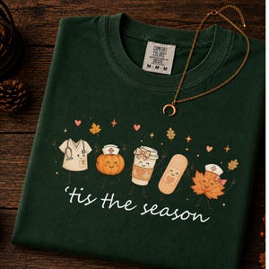 Peut inclure: Un t-shirt vert foncé avec un motif représentant une blouse médicale, une citrouille, un gobelet à café, un pansement et une feuille d'érable, tous avec des visages souriants. Le texte 'tis the season' est imprimé en blanc. Un collier croissant doré.