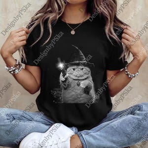 May include: Black t-shirt featuring a grayscale illustration of a cat wearing a witch hat and holding a gun. The cat has a serious expression. The shirt is a casual wear item.