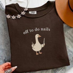 May include: A dark brown t-shirt featuring a graphic of a light brown goose holding a nail polish bottle and a nail file. The text "off to do nails" is printed in white above the goose. The shirt has a crew neckline and a Comfort Colors label.