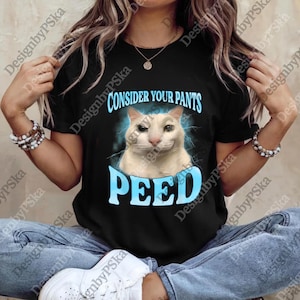 Puede incluir: Camiseta negra con el texto azul "CONSIDER YOUR PANTS PEED". La camiseta presenta un gráfico de gato blanco con una expresión de suficiencia, sobre un efecto de relámpagos azules. Un diseño casual y humorístico.