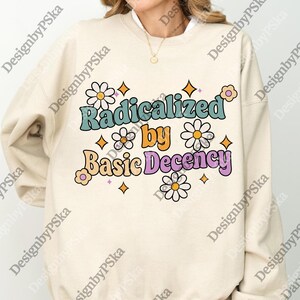 Puede incluir: Sudadera color crema con el texto "Radicalized by Basic Decency" en una fuente retro, adornada con margaritas y estrellas. El diseño presenta una combinación de colores turquesa, naranja y morado, creando una estética vintage. Una prenda casual y cómoda.
