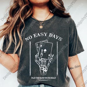 May include: Dark gray t-shirt with the text "NO EASY DAYS" above a graphic of a skeleton hand holding playing cards. The cards show a 2 of hearts and a 7 of spades. The text "PLAY THE HAND YOU'RE DEALT" is below.