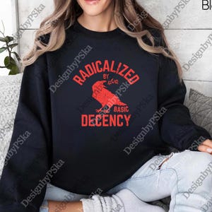 Puede incluir: Sudadera negra con el texto rojo "RADICALIZED BY BASIC DECENCY". Un gráfico de cuervo rojo está centrado debajo del texto. La sudadera es de cuello redondo.