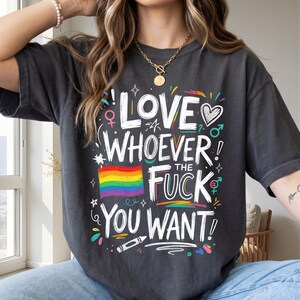 Puede incluir: Una camiseta gris carbón con texto blanco escrito a mano que dice LOVE WHOEVER THE FUCK YOU WANT. El diseño incluye una bandera arcoíris colorida, un corazón y varios símbolos de género en colores brillantes como rosa, azul y amarillo.