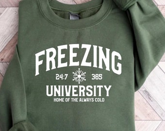 Może przedstawiać: Zielony sweter z białym napisem "Freezing University" z grafiką płatka śniegu i tekstem "24:7 365" oraz "Home of the Always Cold".