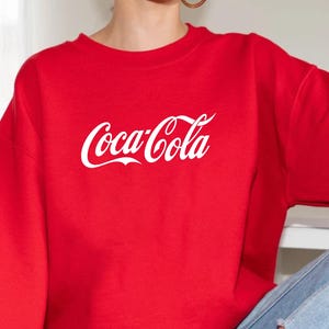 May include: A red crewneck sweatshirt with the Coca-Cola logo in white script across the chest. The sweatshirt is made of a soft material and is paired with blue jeans and white sneakers. The overall style is casual and comfortable.