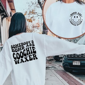 May include: White sweatshirt with black text that reads "Somebody's Bomb Ass Coochie Waxer" on the back and a smiley face with the text "Bomb Ass Coochie Waxer" on the front.