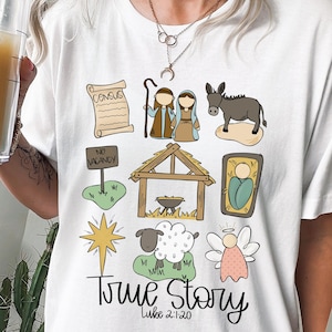 May include: White t-shirt with a colorful illustration of the nativity story. The illustration includes a star, a manger, a donkey, a sheep, an angel, and the text "True Story Luke 2:1-20".