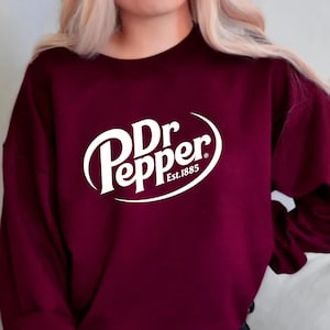 May include: A maroon sweatshirt featuring the Dr Pepper logo in white. The logo includes the brand name in a stylized font and the text "Est.1885". The sweatshirt is a solid color and has a crew neck.
