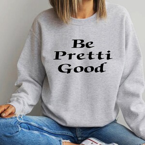 May include: A light gray sweatshirt with the words "Be Pretti Good" printed in black. The sweatshirt is a crew neck style and appears to be made of a soft material. The person in the image is wearing blue jeans.