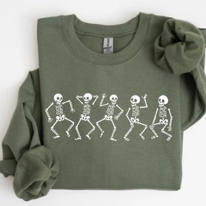 May include: Olive green sweatshirt featuring a white graphic of five dancing skeletons. Each skeleton has the words "FUCK TRUMP" printed on their rib cage. The sweatshirt has a crew neck and long sleeves.