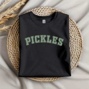 May include: Black t-shirt with a green "PICKLES" graphic in a collegiate font.