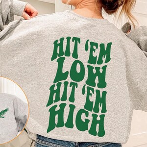 K&ouml;nnte beinhalten: Grauer Pullover mit gr&uuml;ner Grafik, auf dem "Hit 'em Low Hit 'em High" steht.