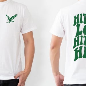 K&ouml;nnte beinhalten: Wei&szlig;es T-Shirt mit einem gr&uuml;nen Adler-Motiv auf der Vorderseite und dem Text "HIT 'EM LOW HIT 'EM HIGH" in Gr&uuml;n auf der R&uuml;ckseite.