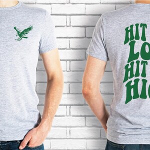 K&ouml;nnte beinhalten: Ein hellgraues T-Shirt mit einem gr&uuml;nen Vogelmotiv auf der Vorderseite und dem Text "Hit 'em Low Hit 'em High" in Gr&uuml;n auf der R&uuml;ckseite.