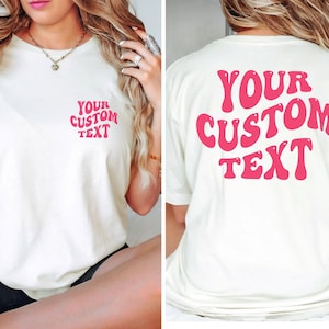 May include: White t-shirt with pink retro font that says "YOUR CUSTOM TEXT" on the front and back.