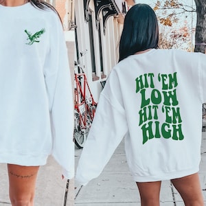 K&ouml;nnte beinhalten: Zwei wei&szlig;e Sweatshirts mit gr&uuml;nem Text und Grafiken. Ein Sweatshirt hat eine gr&uuml;ne Adlergrafik auf der Vorderseite. Das andere Sweatshirt hat den Text "Hit 'em Low Hit 'em High" auf der R&uuml;ckseite.