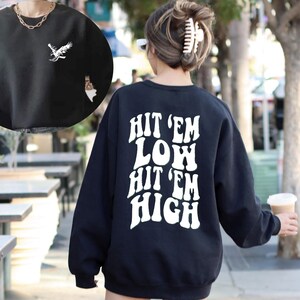 K&ouml;nnte beinhalten: Schwarzer Pullover mit wei&szlig;em Text "Hit 'em Low Hit 'em High" auf der R&uuml;ckseite. Der Pullover hat eine lockere Passform und lange &Auml;rmel.